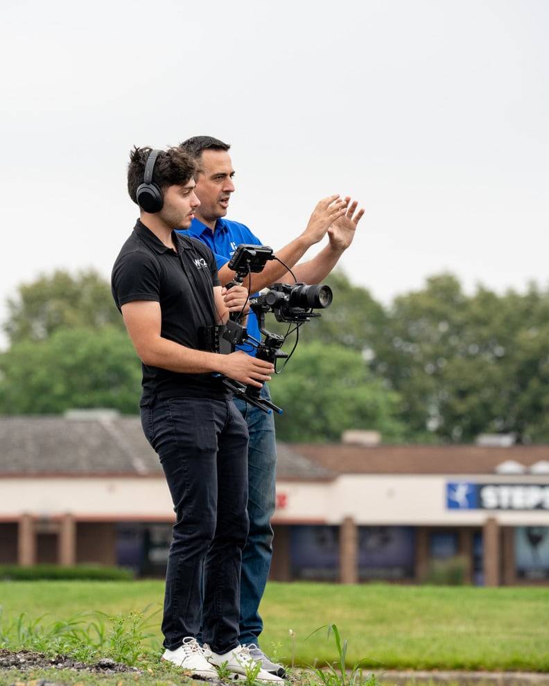 Camera operator with headphones holding a gimbal rig beside a man in a blue polo during an outdoor video shoot
