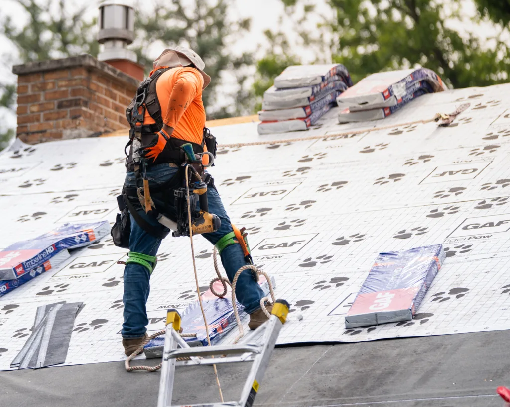 Roofer in an orange shirt and safety harness standing on GAF underlayment with stacked shingle bundles