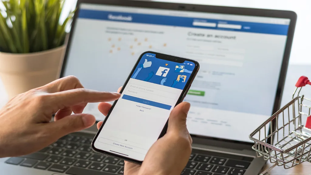 Hands holding an iPhone with the Facebook login screen in front of a laptop showing a Facebook signup page