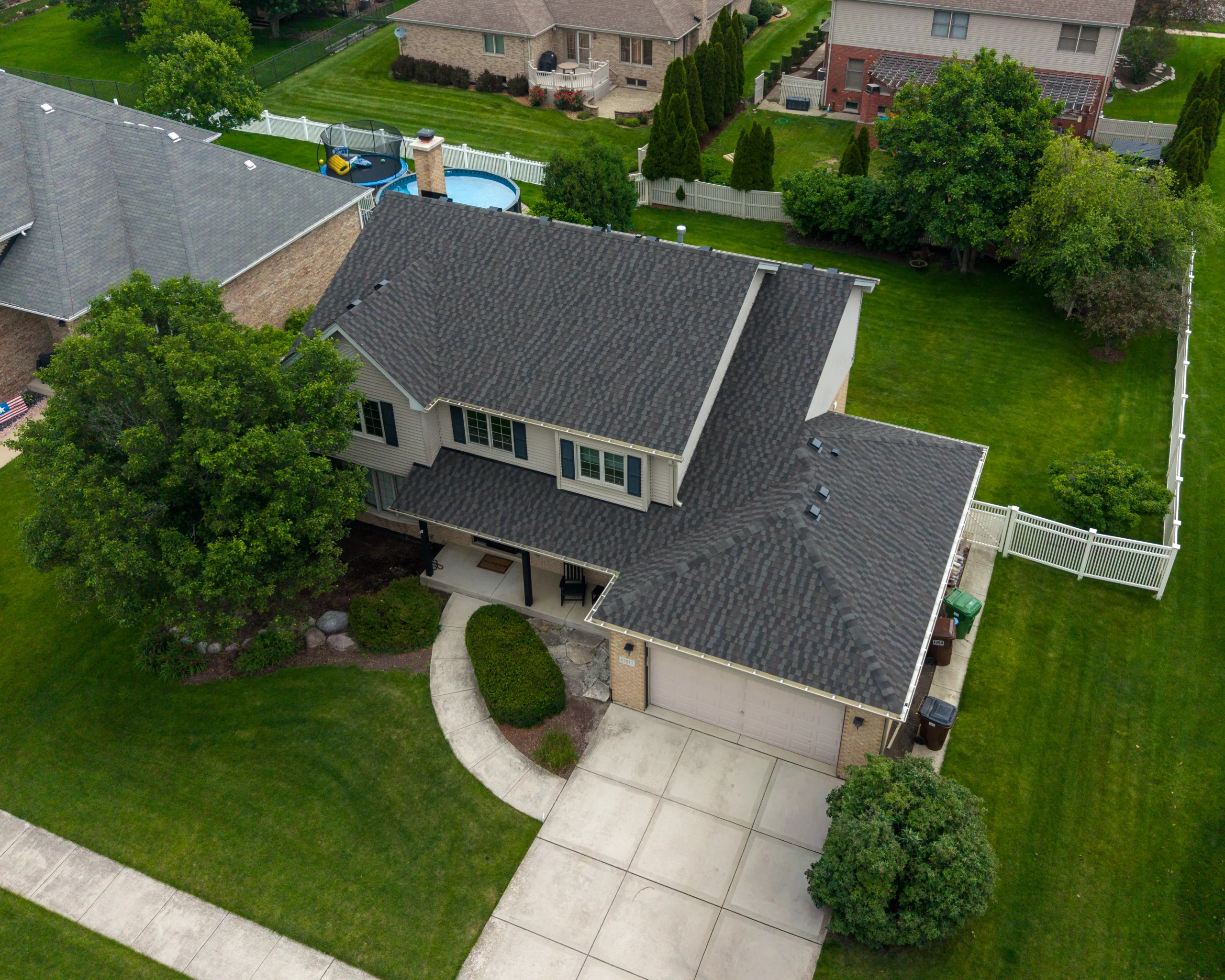 Aerial drone view of a two-story suburban home with a gray architectural shingle roof, driveway, and fenced yard