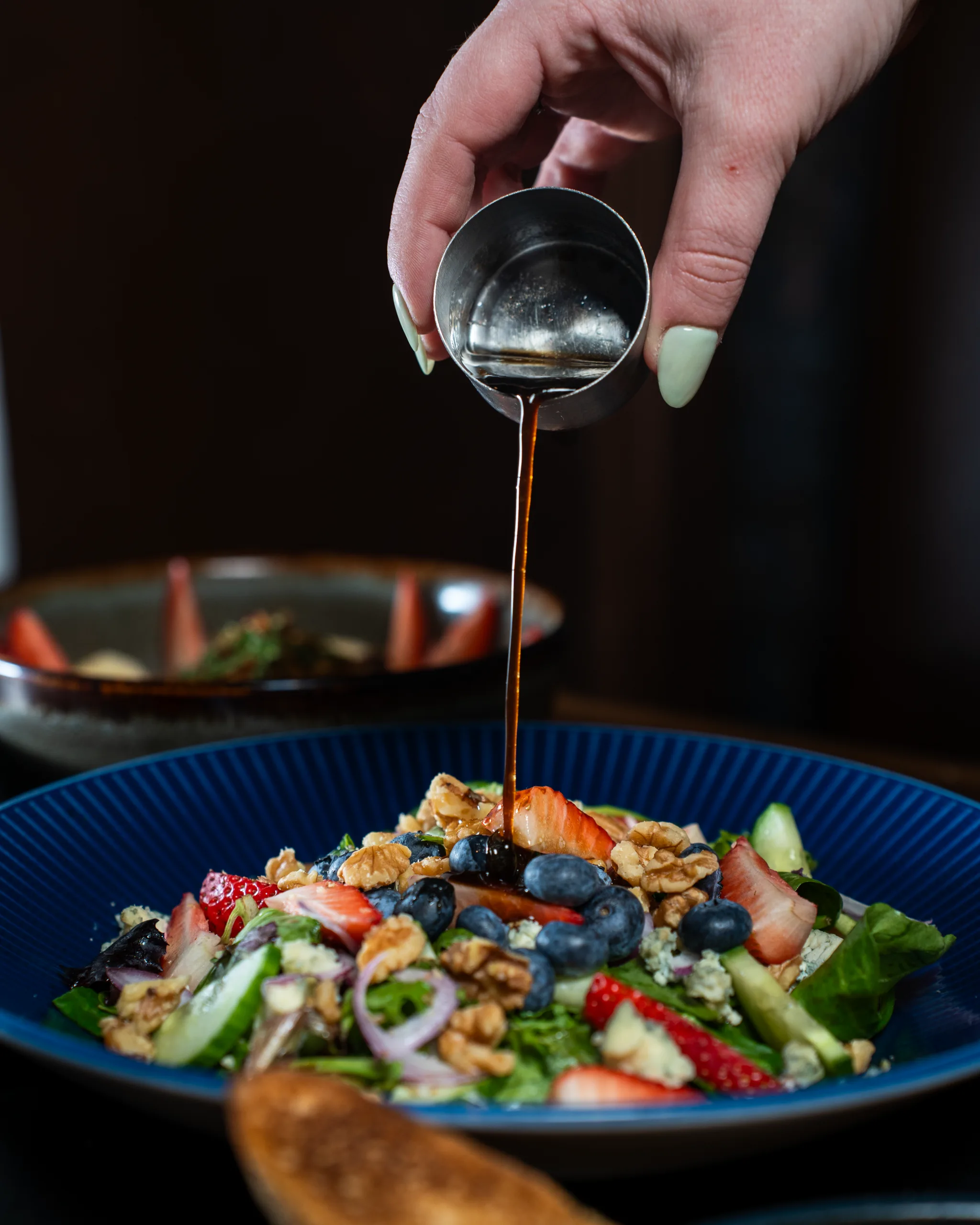 Hand pouring balsamic dressing from a steel cup onto a berry and walnut salad on a blue plate