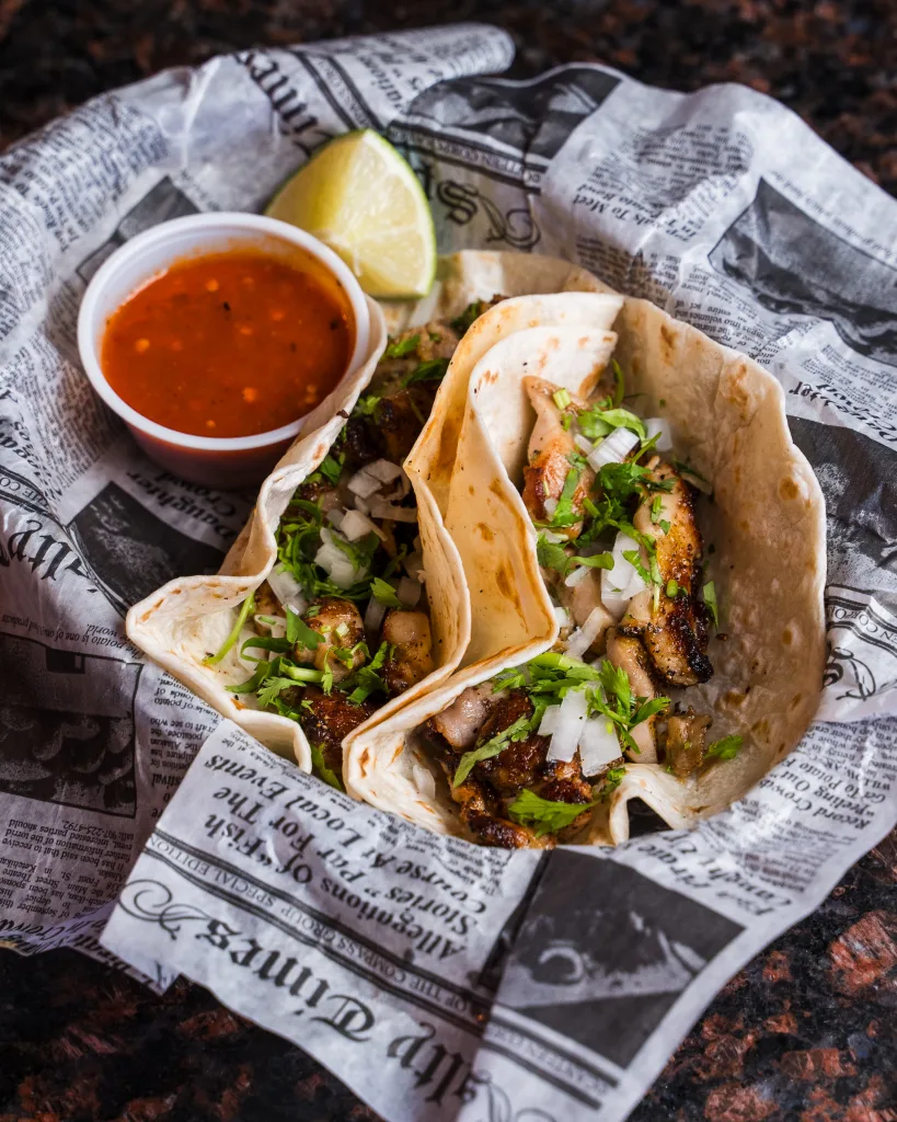 Two grilled chicken tacos topped with onion and cilantro in a newspaper-lined basket with salsa and lime