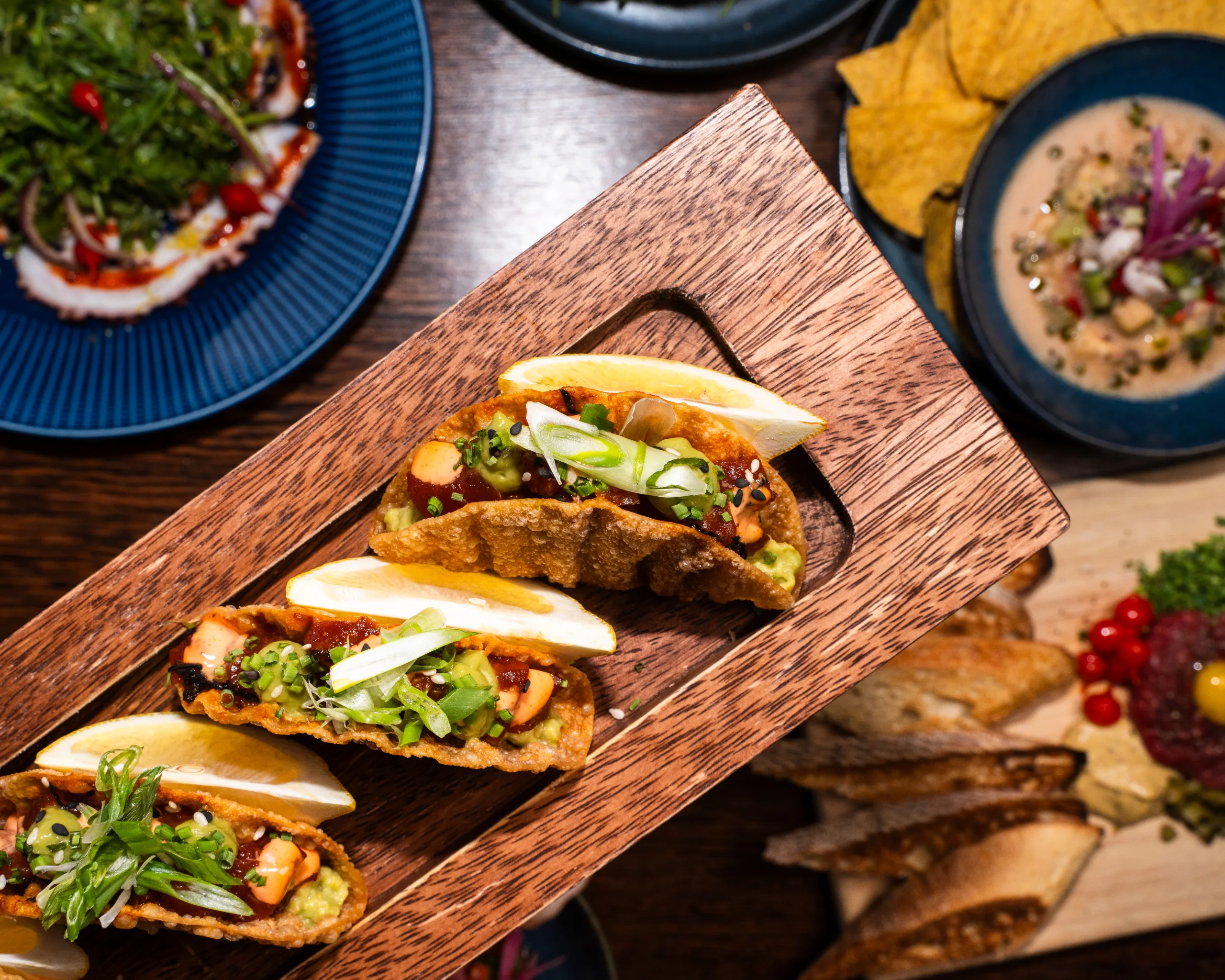 Three crispy tacos with guacamole, scallions, and lemon wedges on a wooden board beside ceviche and tortilla chips