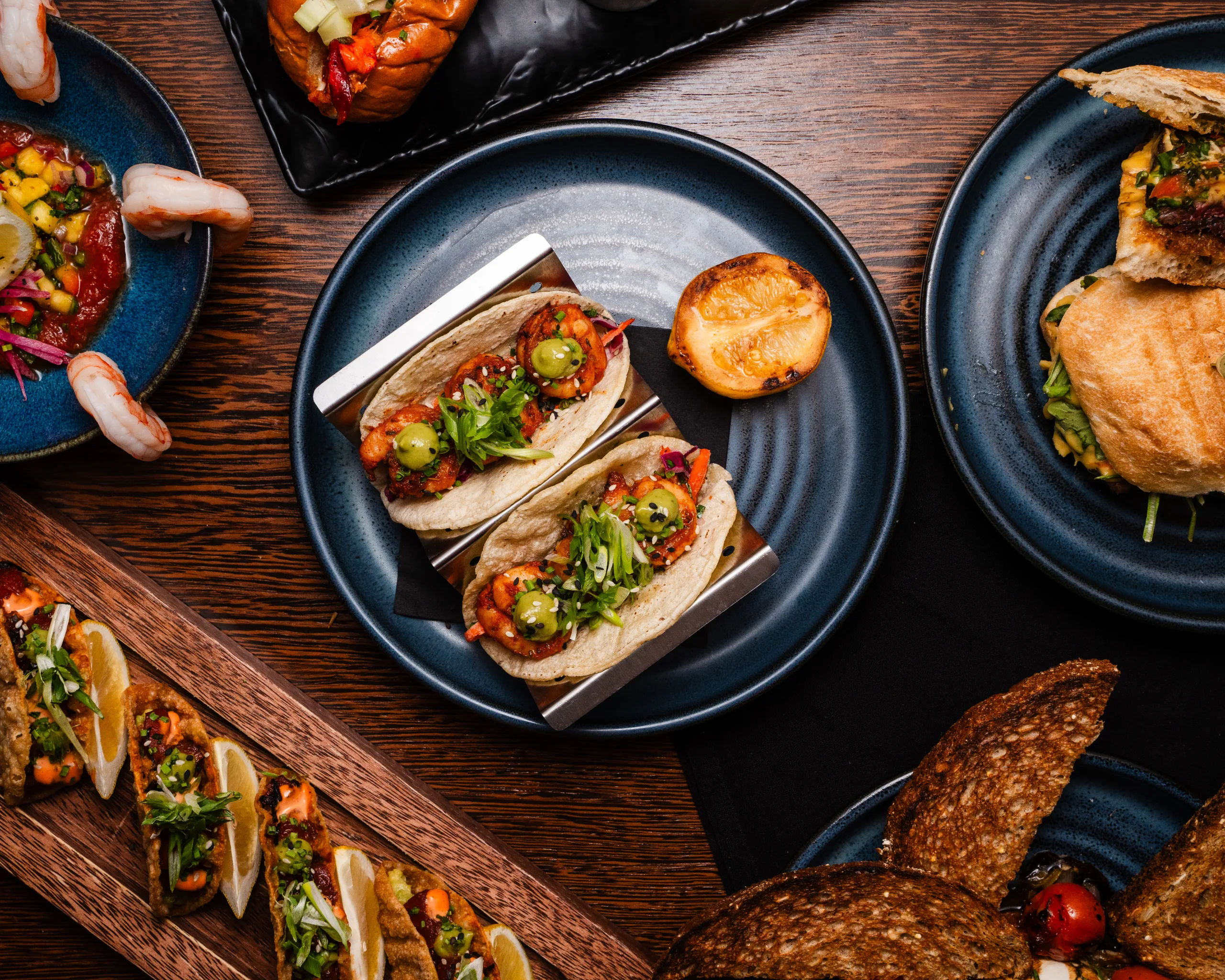 Overhead spread of shrimp tacos with charred lemon, crispy wonton tacos, shrimp cocktail, and sandwiches