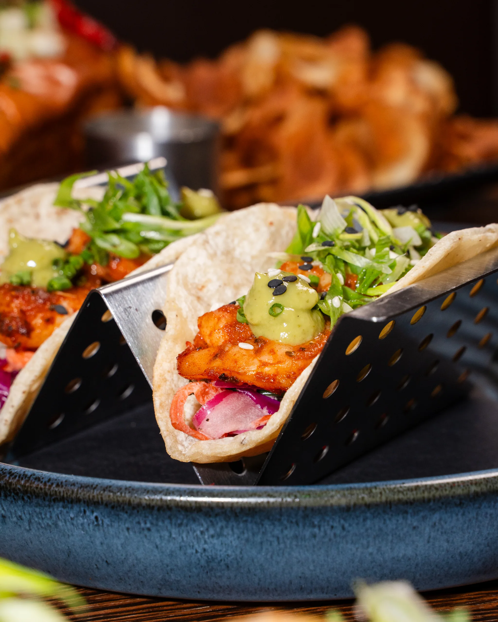 Shrimp tacos topped with guacamole, green onion, and sesame seeds in a metal stand on a blue ceramic plate