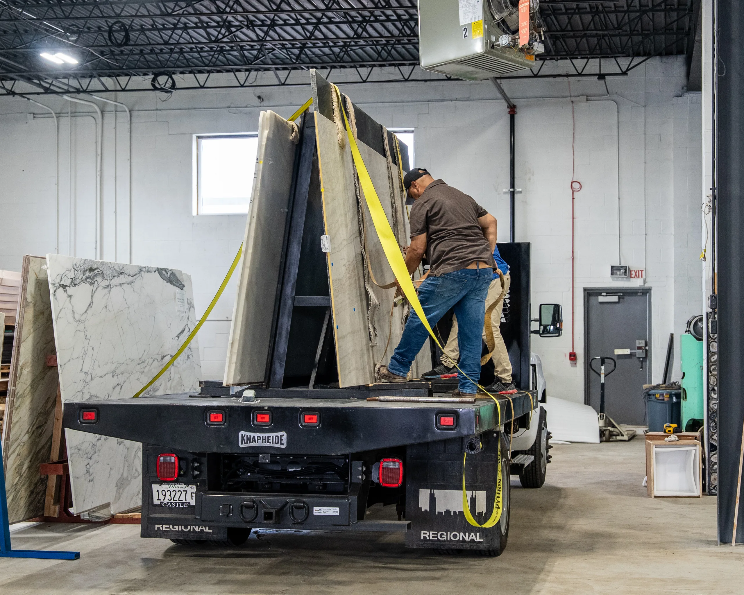 Workers strapping marble slabs upright on a flatbed truck inside a stone warehouse