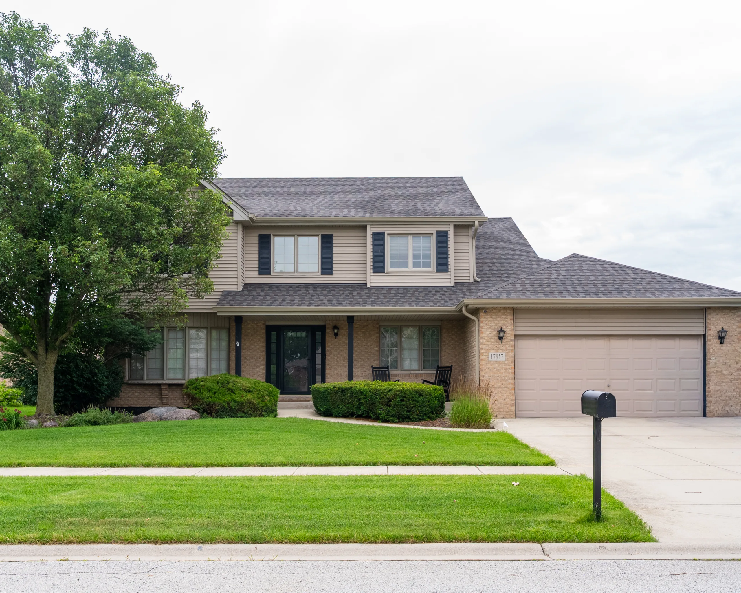 Street view of a two-story home with tan siding, gray asphalt shingle roof, landscaped lawn, and black mailbox
