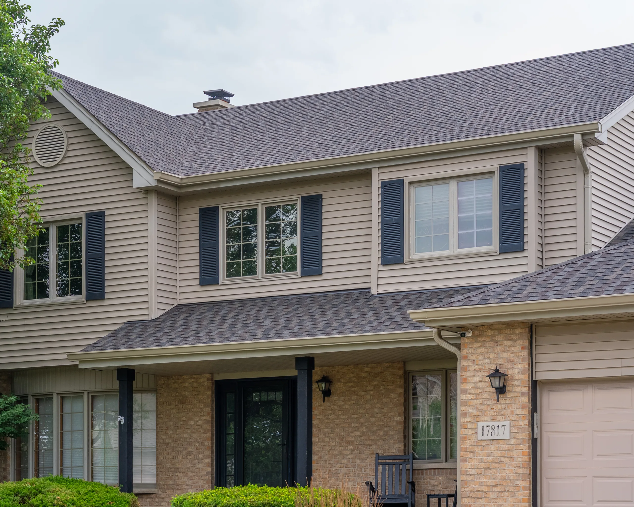Front facade of a two-story home with gray asphalt shingles, tan siding, black shutters, and brick entryway
