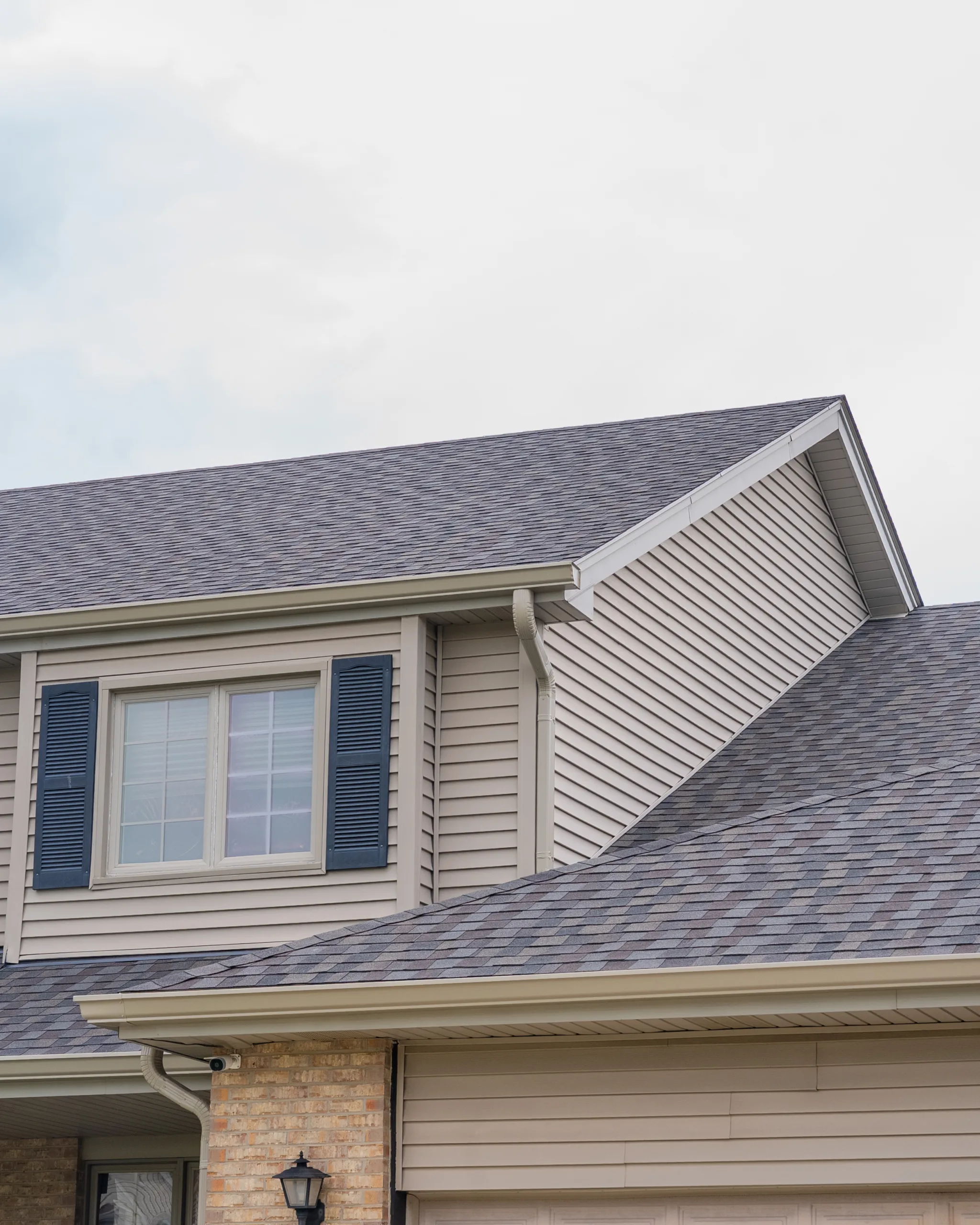 Roofline detail of multi-tone asphalt shingles meeting a gabled dormer with tan siding and black shutters