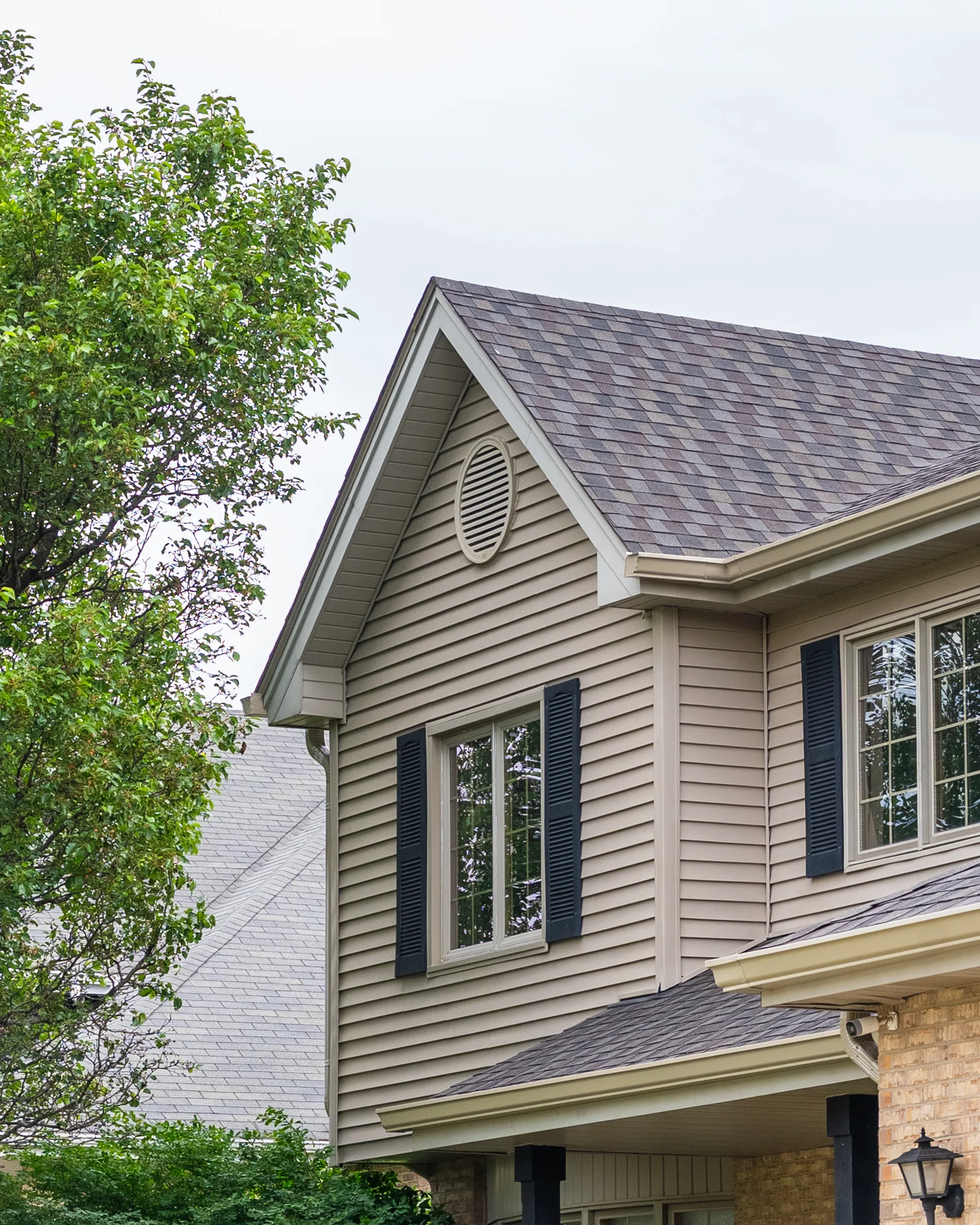 Gable end of a two-story home with tan siding, oval vent, black shutters, and multi-tone asphalt shingles