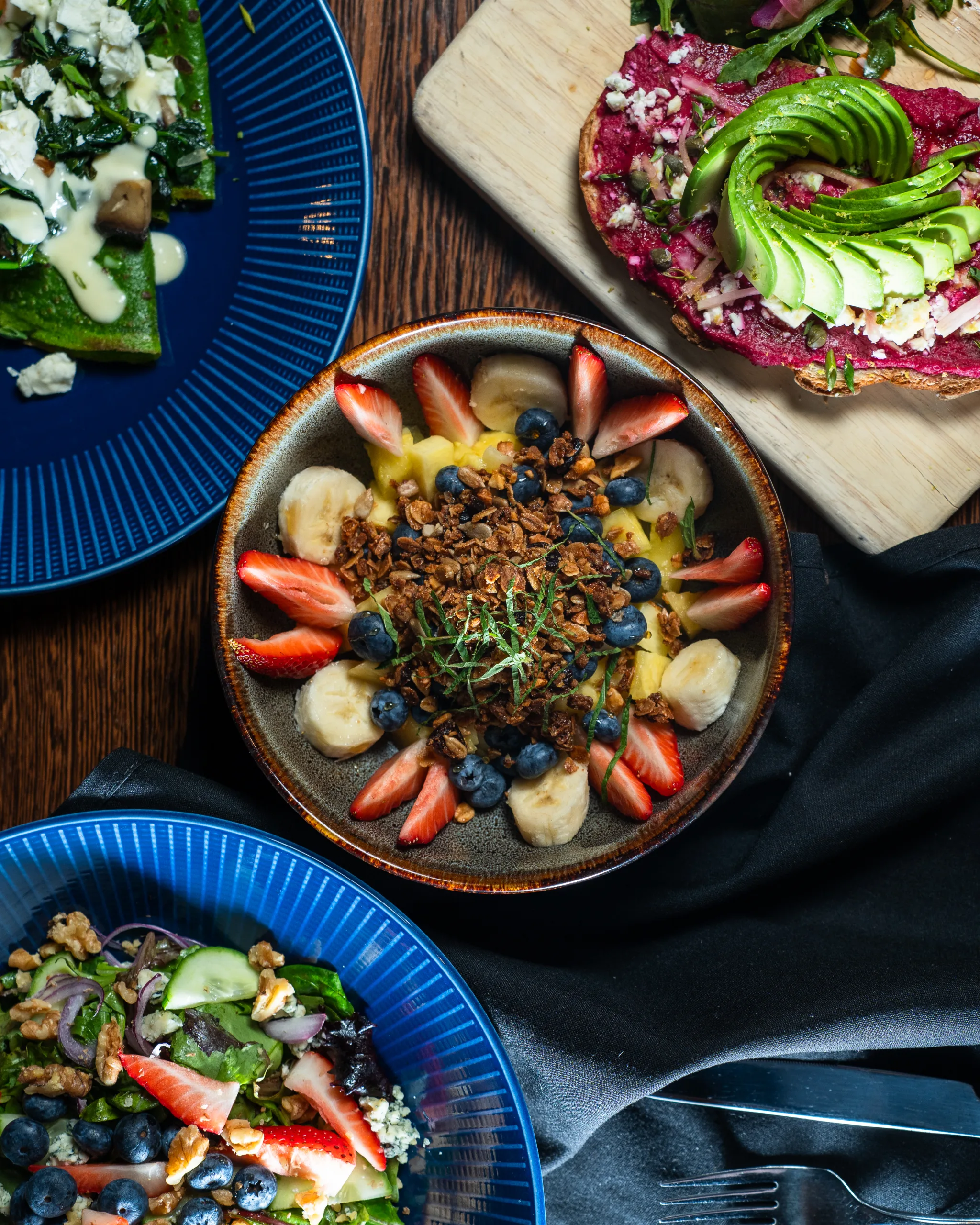 Overhead view of a fruit and granola bowl, berry salad, avocado beet toast, and spinach crepe on a wood table