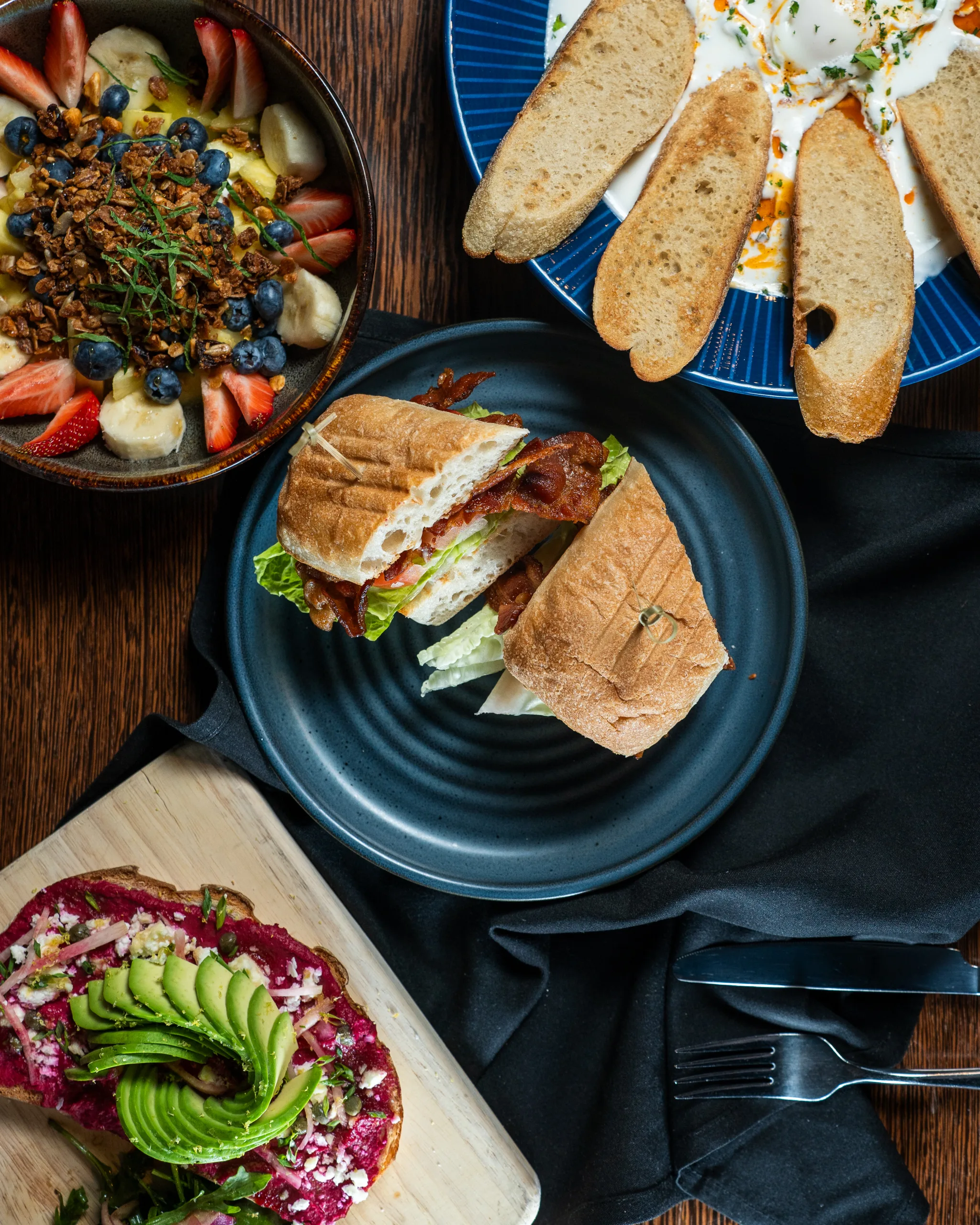 Overhead view of a bacon sandwich on ciabatta, fruit granola bowl, avocado beet toast, and toasted bread with dip
