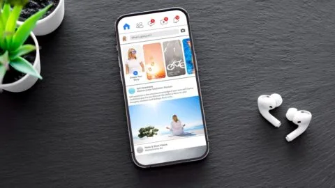 Smartphone on a dark slate surface displaying a Facebook feed, next to wireless earbuds and a succulent