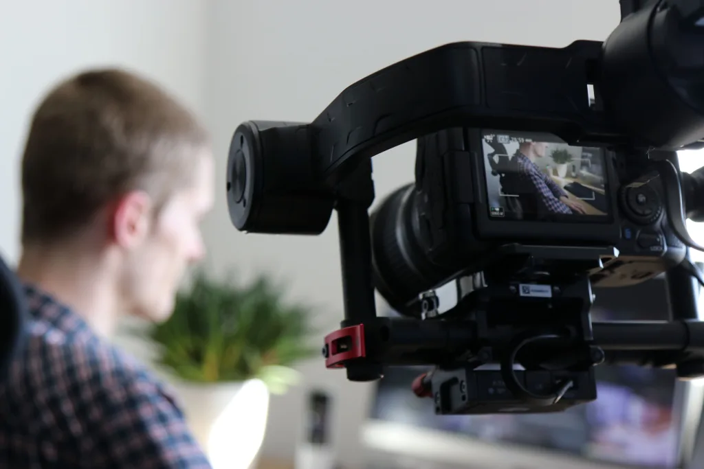 Videographer operating a gimbal-mounted camera, its flip screen showing a man working at a desk
