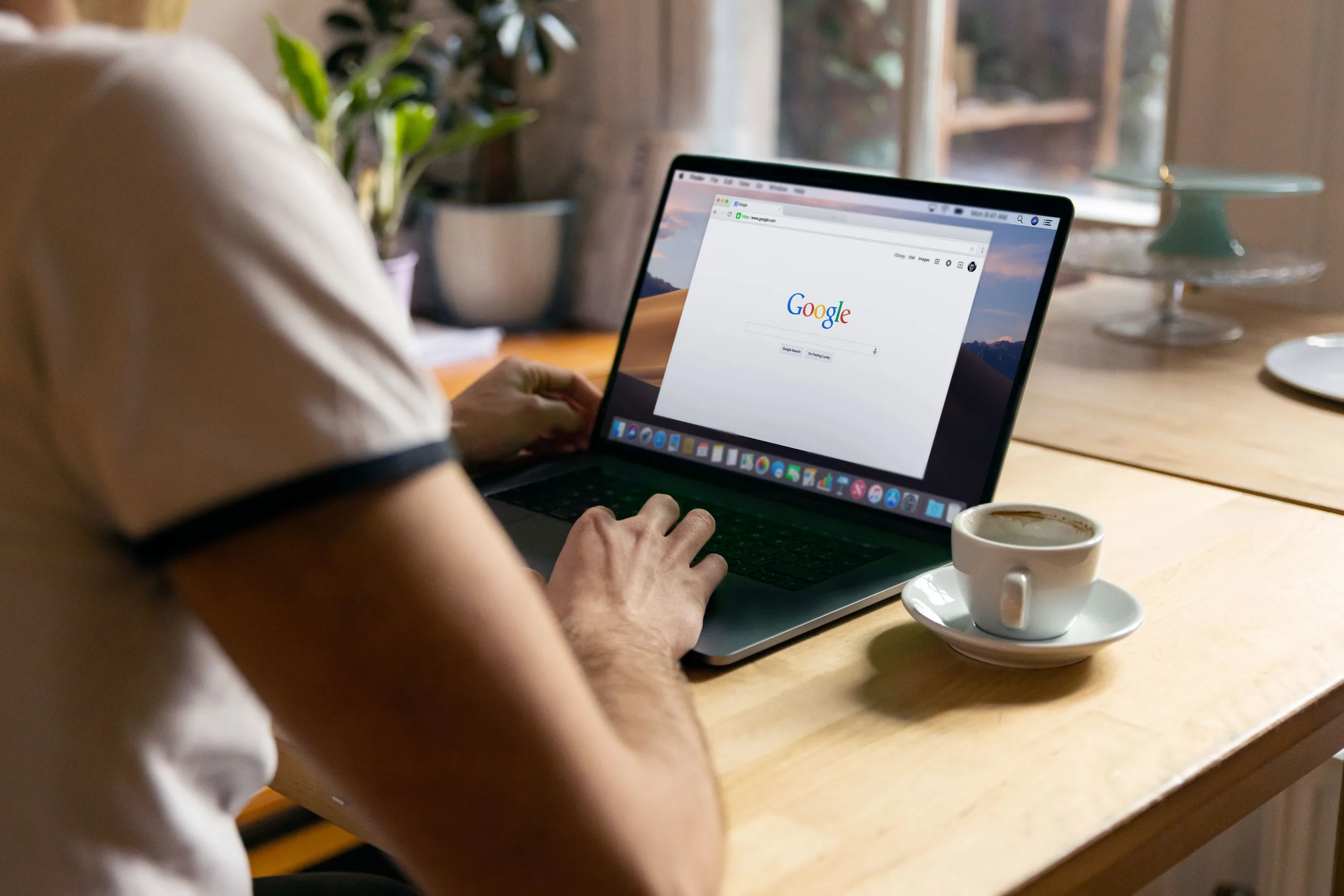 Man typing on a MacBook with the Google homepage on screen, coffee cup on a wooden table beside him