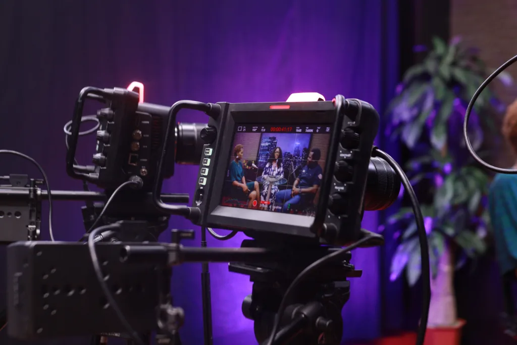 Cinema camera monitor framing three people during a talk show interview on a purple-lit set