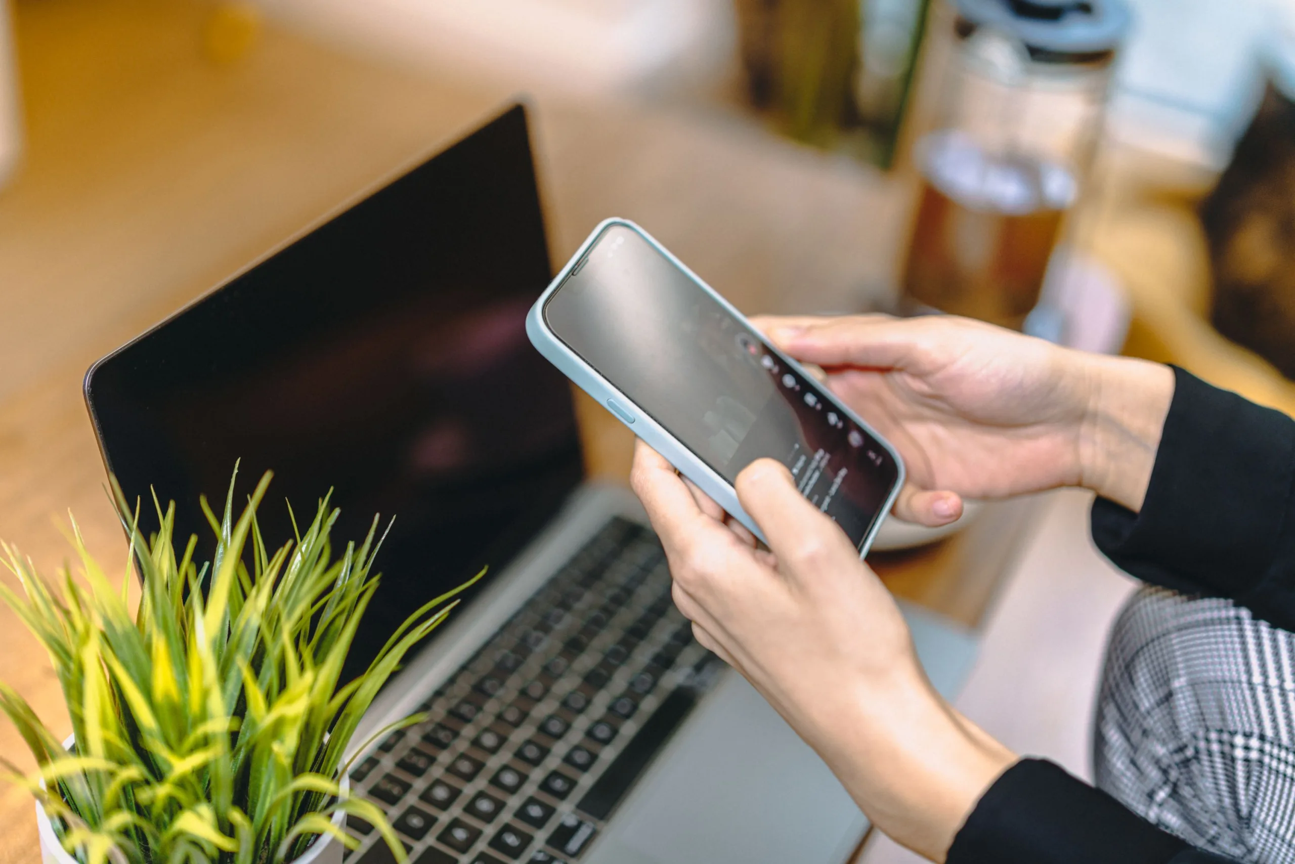 Person holding a smartphone with both hands at a desk in front of an open laptop and a potted plant