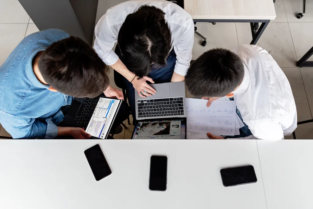 Overhead view of three people collaborating at a desk with laptops, handwritten notes, and smartphones