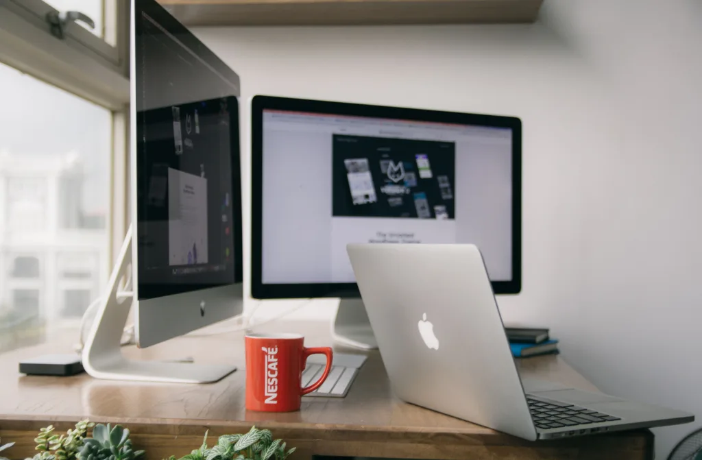 Home office desk with an iMac, external monitor, open MacBook, red Nescafe mug, and plants by a window