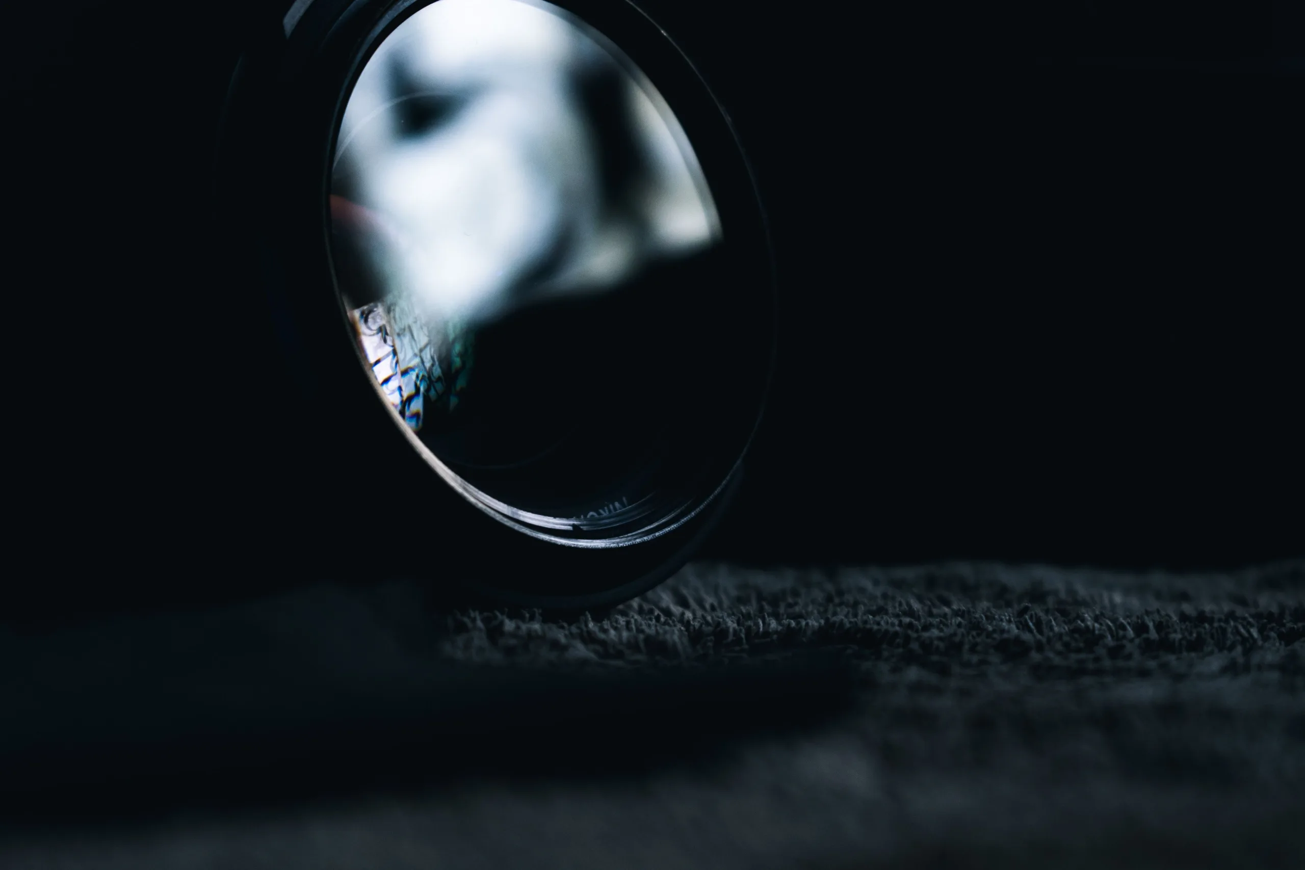 Camera lens resting on dark fabric in low light, with a blurred reflection on the front glass element