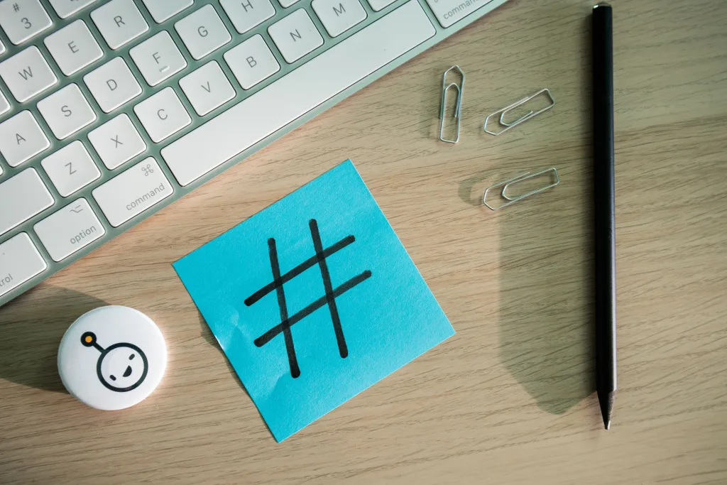 Blue sticky note with a hand-drawn hashtag beside a keyboard, paper clips, a pencil, and a robot pin