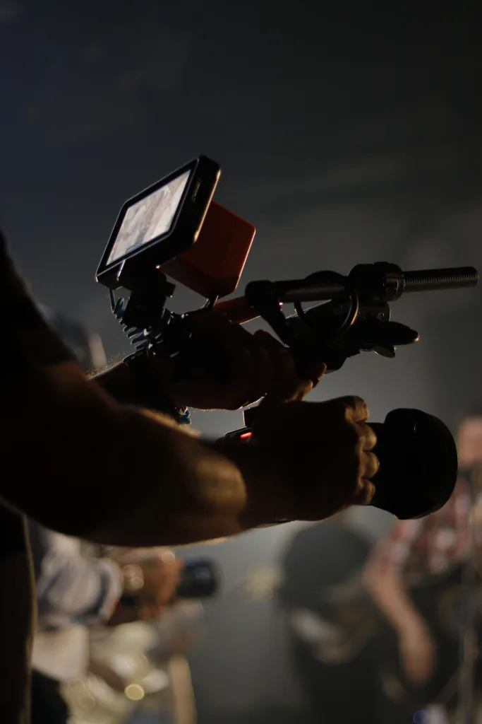 Camera operator's hands on a handheld video rig with monitor and shotgun mic in low light