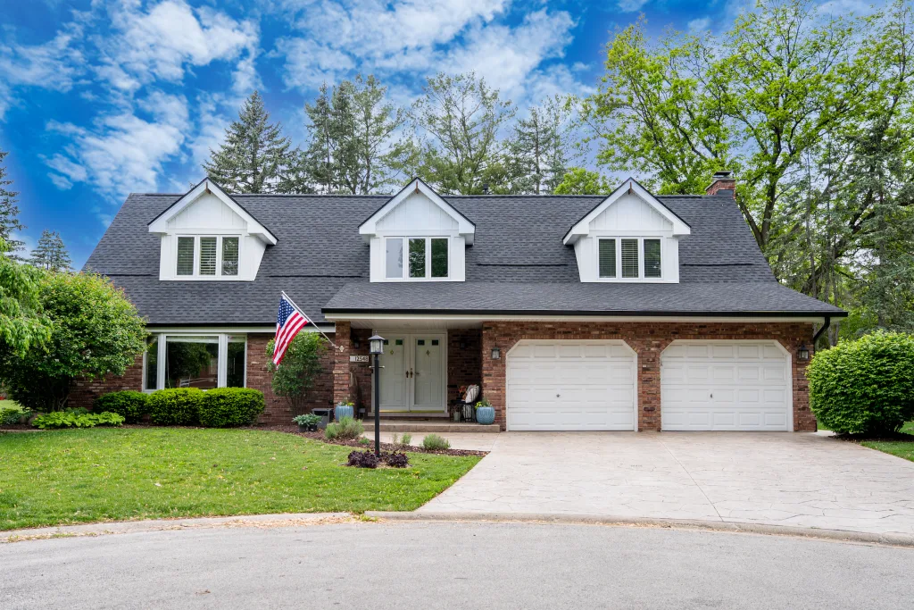 Two-story brick home with three white dormers, dark asphalt shingle roof, American flag, and two garage doors